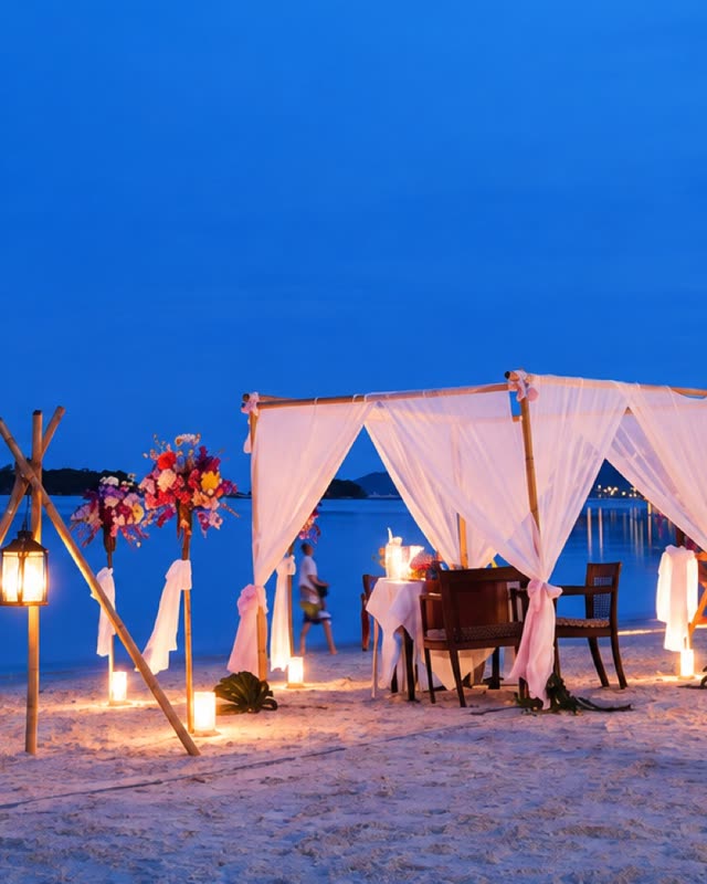 Candlelit canopy dinner table set on the beach at dusk, lanterns and flowers either side