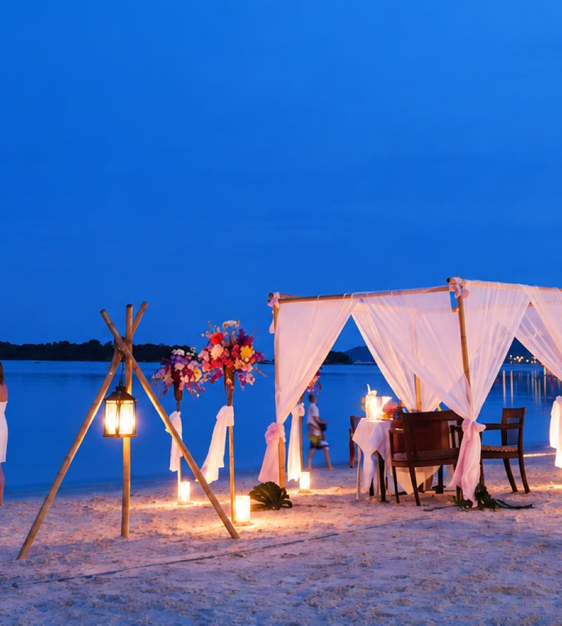 Candlelit canopy dinner table set on the beach at dusk, lanterns and flowers either side