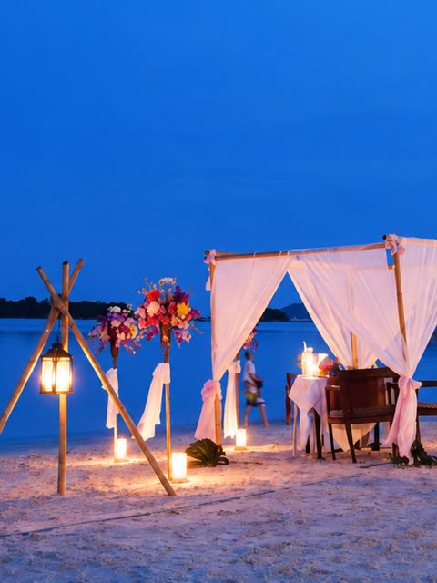 Candlelit canopy dinner table set on the beach at dusk, lanterns and flowers either side