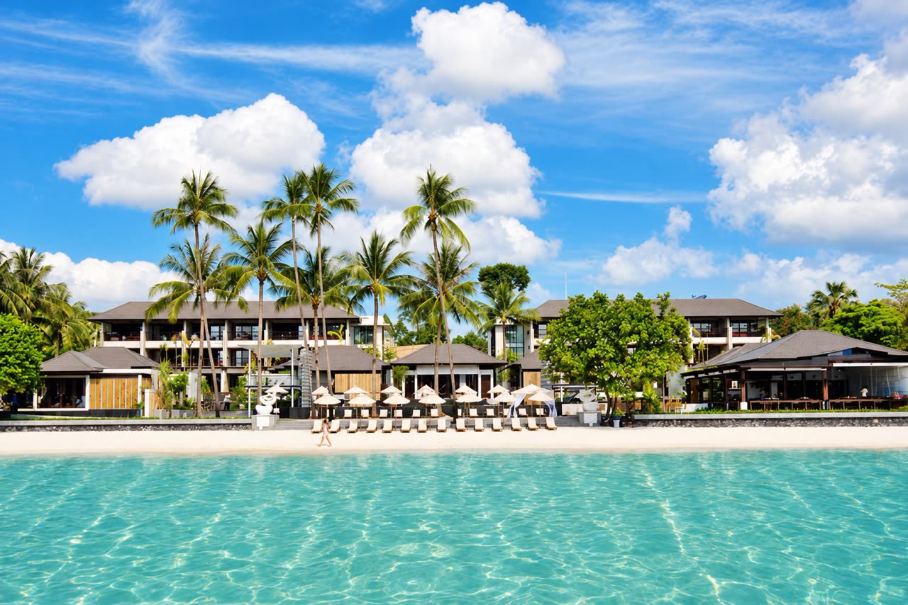 Palm trees and low timber-roofed resort buildings behind a white beach and turquoise water