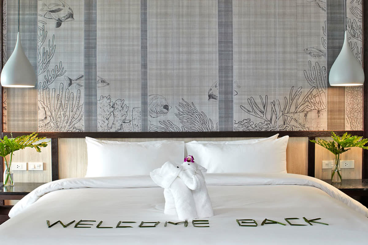 King bed with a folded-towel elephant and a “Welcome back” message beneath a wall panel with fish and coral drawings